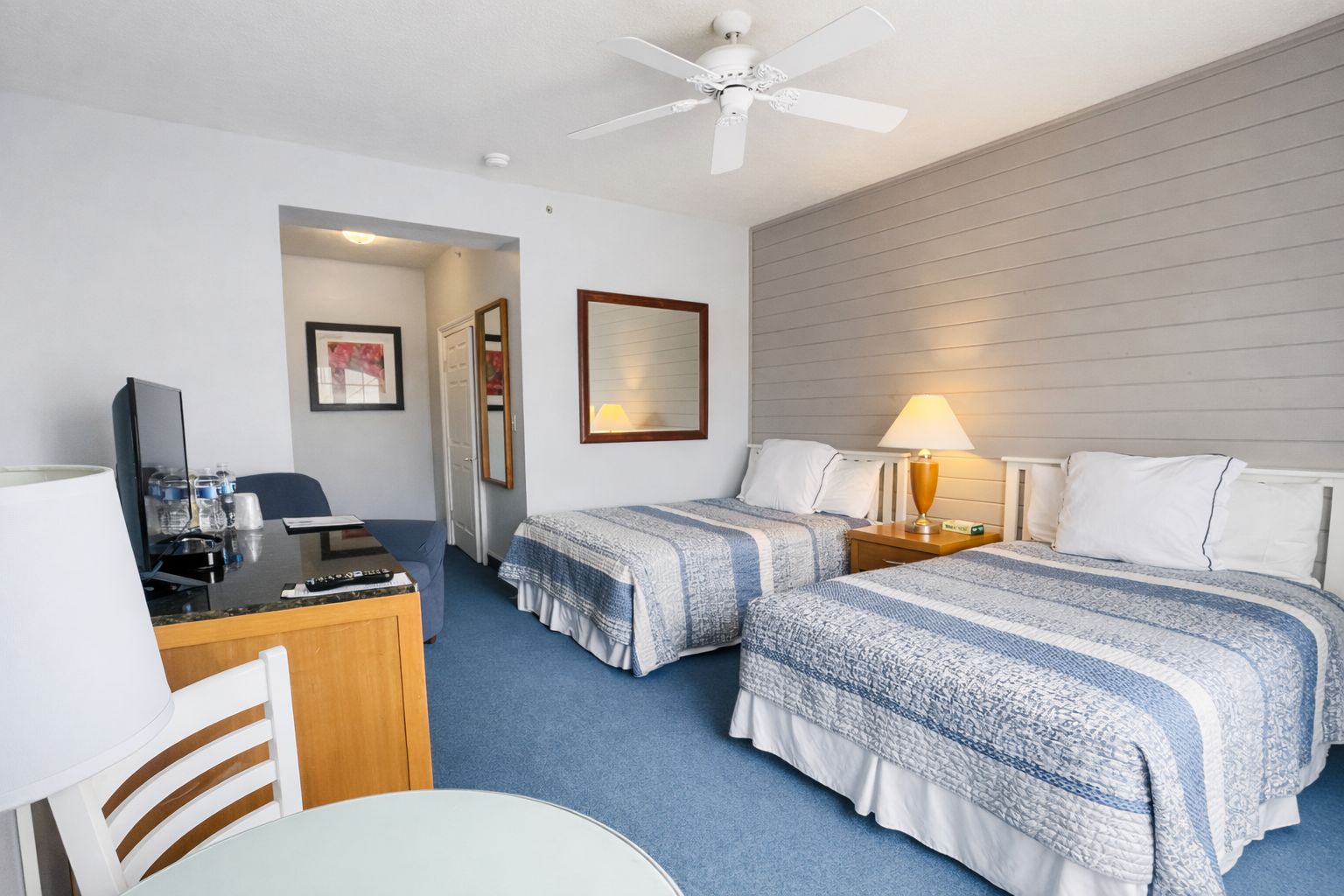 Quarto de hotel com duas camas, uma escrivaninha, uma cadeira, um ventilador de teto e um quadro pendurado na parede.