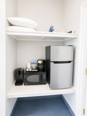 A kitchenette with a refrigerator, microwave, coffee maker, and a shelf with bottles and a kettle.