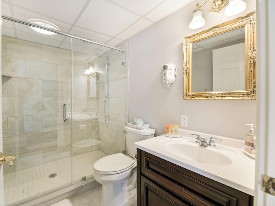 Une salle de bain avec une douche à parois de verre, des toilettes blanches, un meuble-lavabo avec un miroir et des articles de toilette posés sur le lavabo.