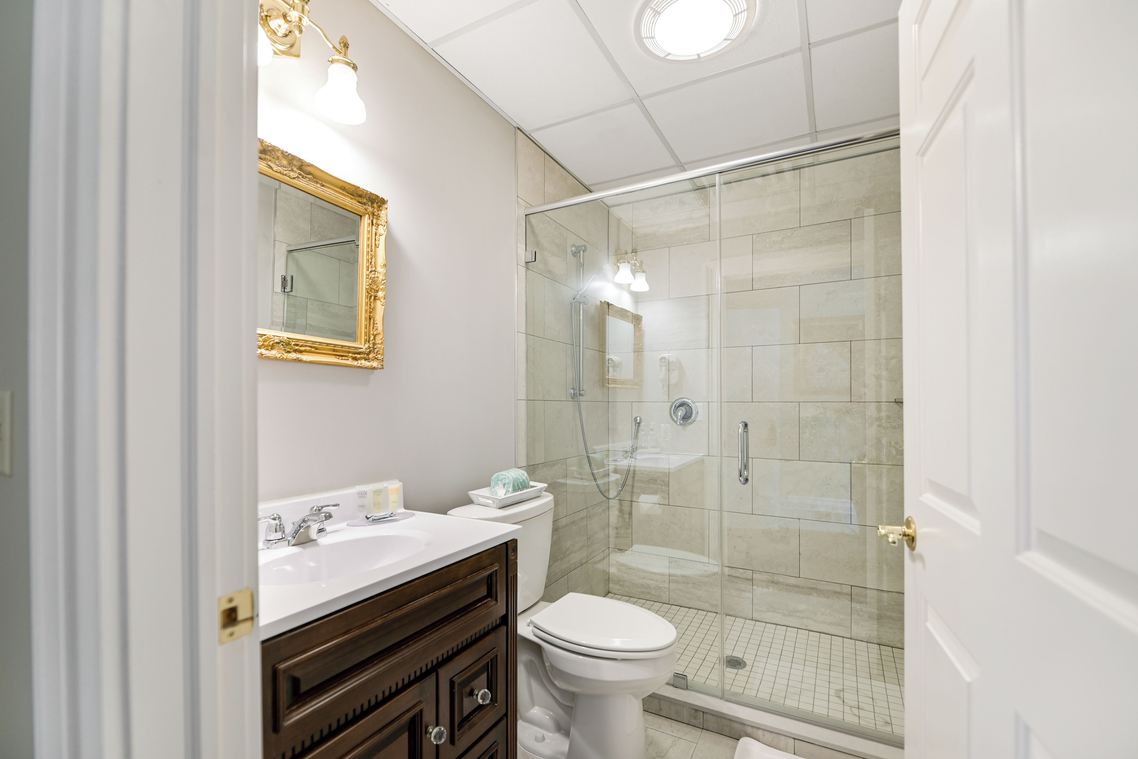 A bathroom with a white vanity, a toilet, a glass shower enclosure, and a mirror with a gold frame.