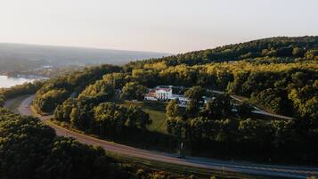 A large estate with a main building surrounded by dense forest, a road curving through the landscape, and a body of water in the distance.