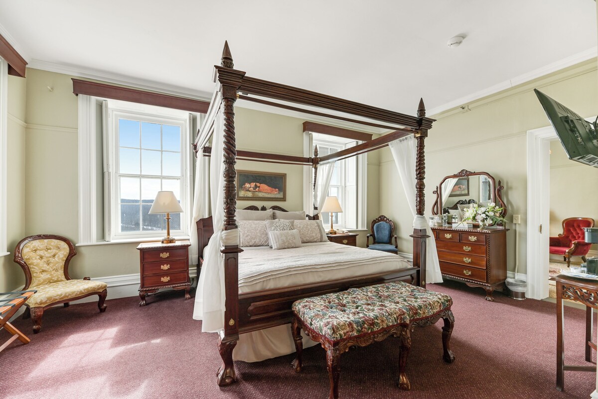 A traditional four-poster bed with a canopy, a sitting bench, and a wooden dresser with a mirror.