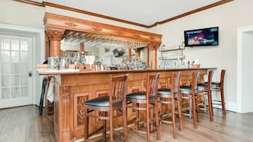 Una zona de bar con taburetes de madera, una barra con botellas y vasos, y un televisor montado en la pared.