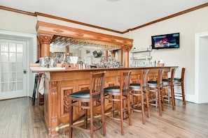 Una zona de bar con taburetes de madera, una barra con botellas y vasos, y un televisor montado en la pared.