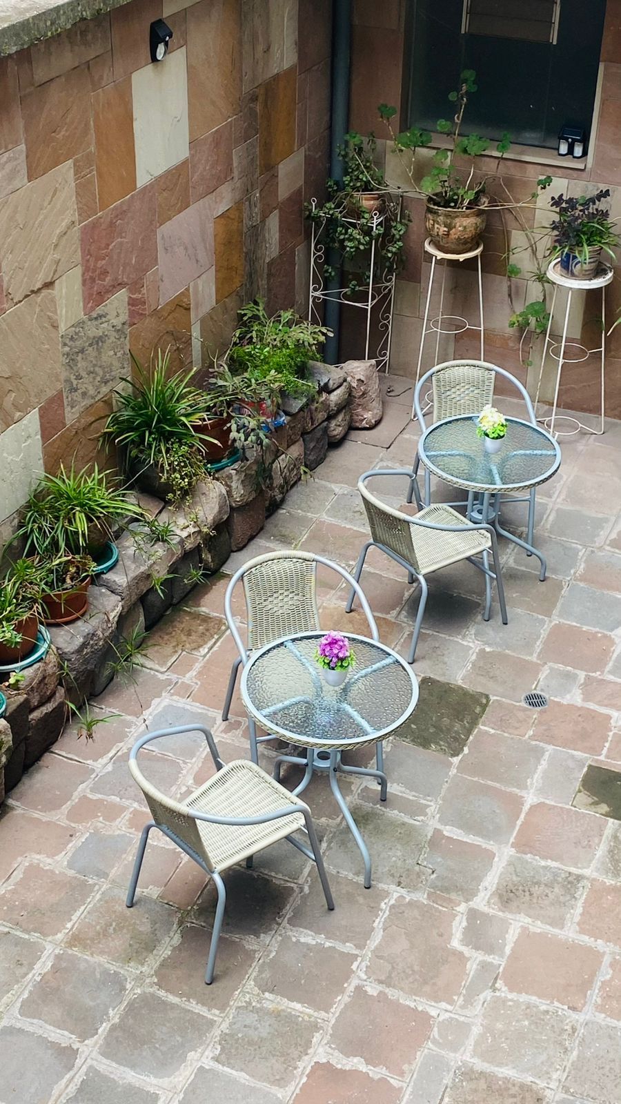 Une terrasse avec un sol en pierre, des chaises et des tables en métal, entourée de plantes en pot.