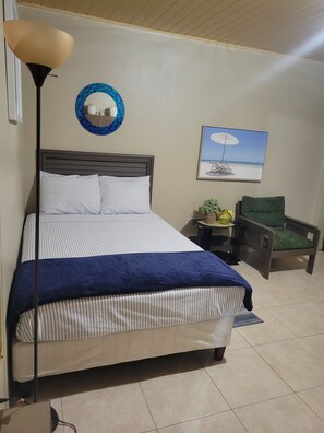 Un dormitorio con cama, mesita de noche, silla, lámpara, una obra de arte en la pared con una escena de playa y un espejo decorativo.
