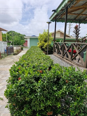 A pathway lined with dense green shrubbery, leading to a covered outdoor seating area with a metal railing.