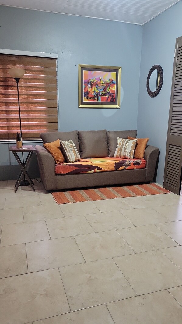A living room with a sofa, a painting, a lamp, and a window with blinds.