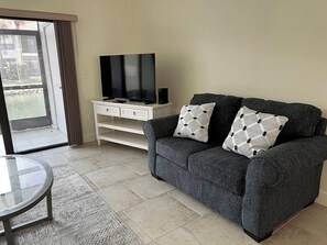 Ein Wohnzimmer mit einer Couch, einem Glastisch, einem Fernseher auf einem weißen Schrank und einer Glastür, die zu einem Außenbereich führt.