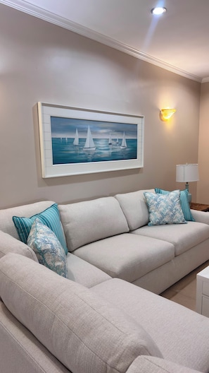 Un salon avec un canapé beige, des coussins à motifs, un tableau encadré représentant des voiliers, une lampe fixée au mur et une petite table avec une lampe.
