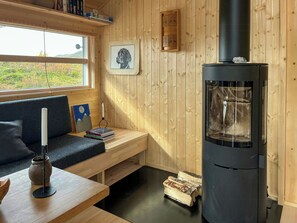 Una habitación acogedora con interior de madera, un estufa de leña negra, una ventana con vistas al verdor, un cuadro en la pared y un banco con libros y una vela.