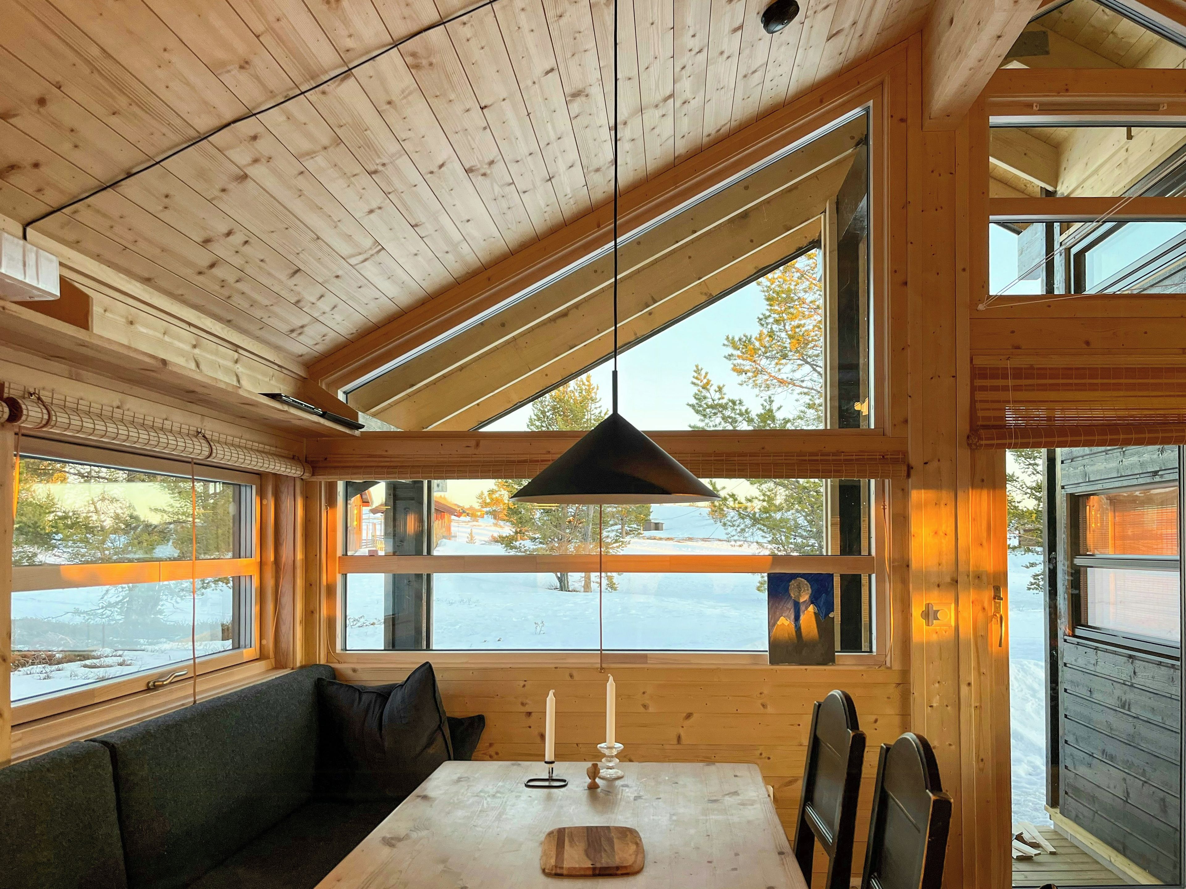 A cabin interior with a wooden ceiling, large windows, a table with a candle, and a bench.