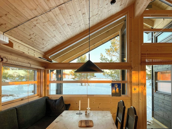 A cabin interior with a wooden ceiling, large windows, a table with a candle, and a bench.