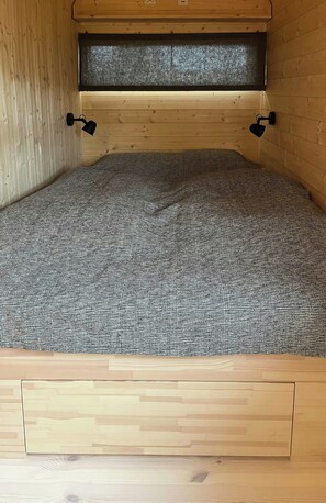 Una cama con colcha estampada en una habitación de madera con cabecera de listones y una pequeña repisa arriba.