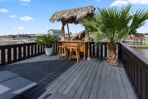 Una terraza de madera con un bar tiki de techo de paja, una palmera y vista al agua.