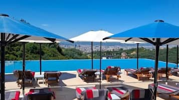 A rooftop lounge area with sun loungers and umbrellas, overlooking a cityscape and a body of water.
