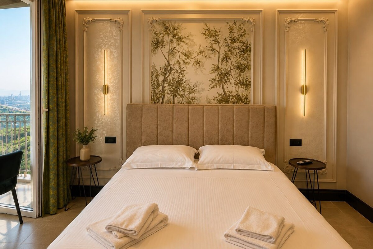 A hotel room with a large bed, two bedside tables, a view of the city, and a decorative wall with a tree mural.