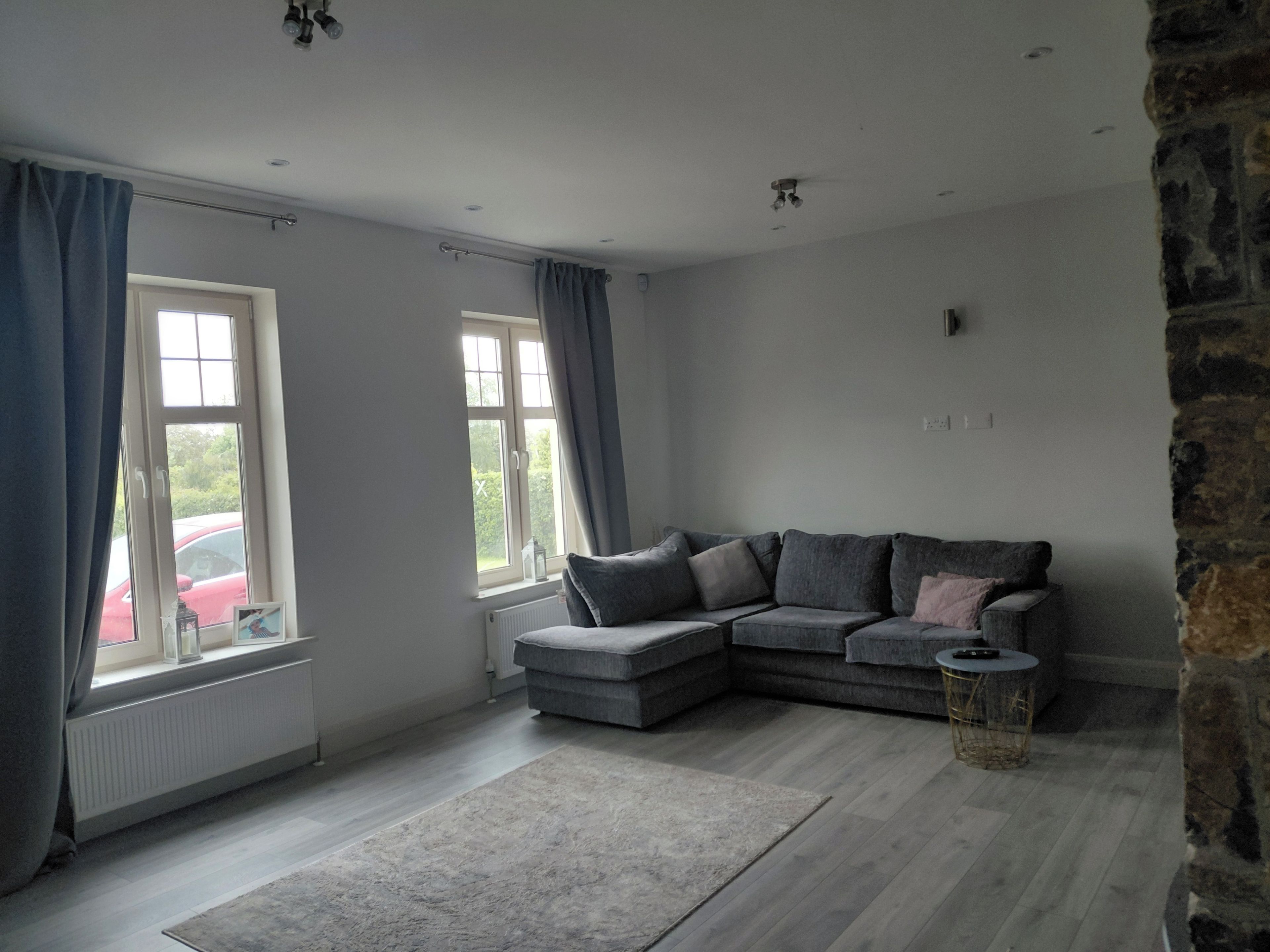 A modern living room with a grey sectional sofa, a small round table, and a grey rug. There are two windows with curtains, a radiator, and a stone wall.