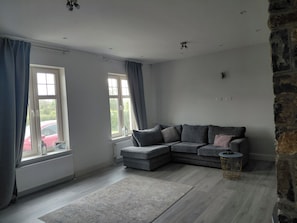 A modern living room with a grey sectional sofa, a small round table, and a grey rug. There are two windows with curtains, a radiator, and a stone wall. - Limerick Luxe Villa Retreat (Limerick)