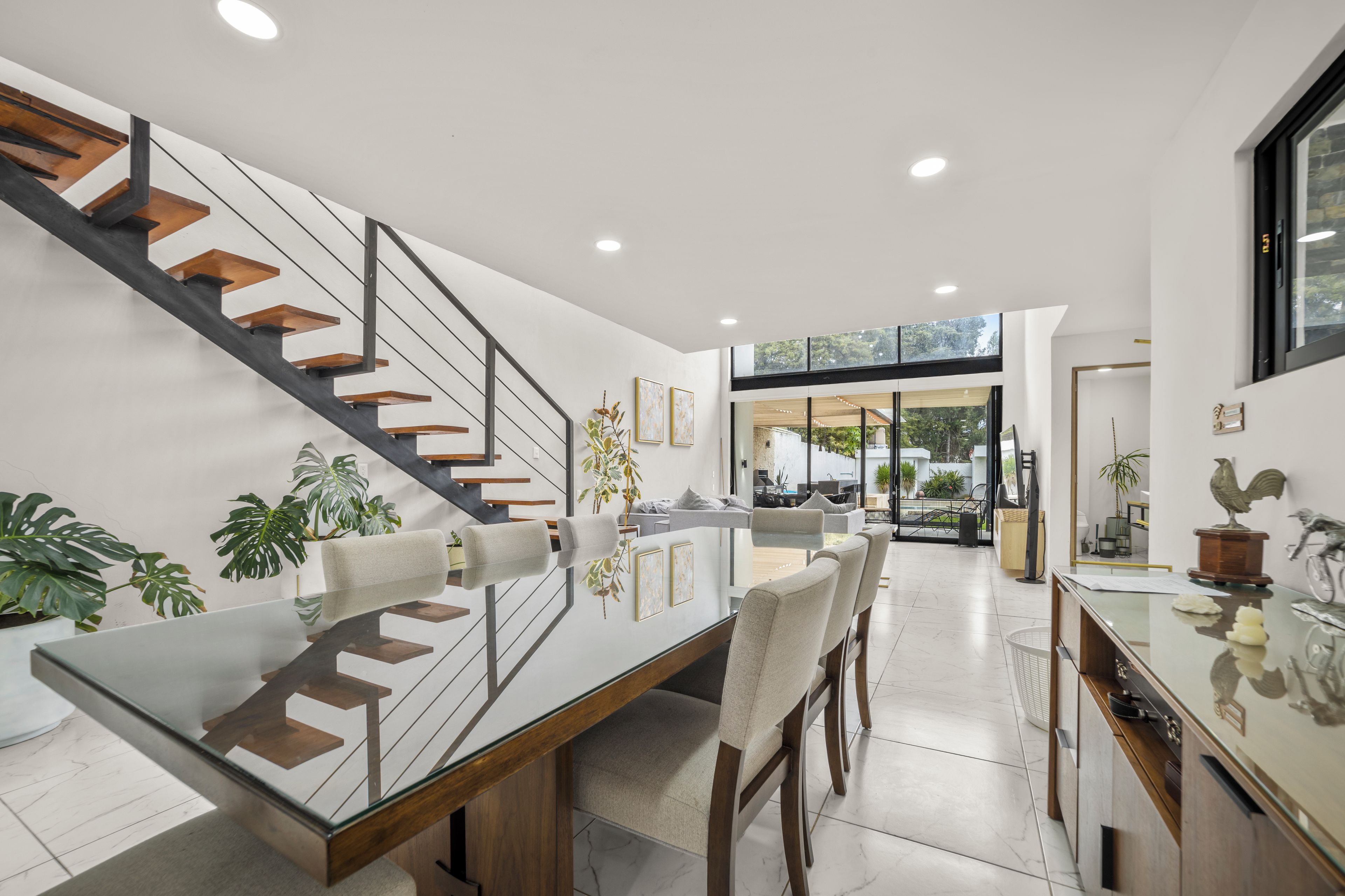 A modern interior with a glass dining table, wooden chairs, a staircase with metal railings, and a view of the outdoors through large windows.