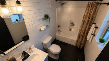 Een moderne badkamer met een wit toilet, een badkuip en een douche. Twee wandlampen, een spiegel en een plank met planten.