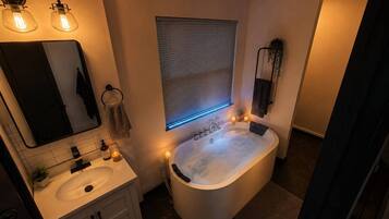 A bathroom with a freestanding bathtub, a sink, a mirror, and a window with blinds.