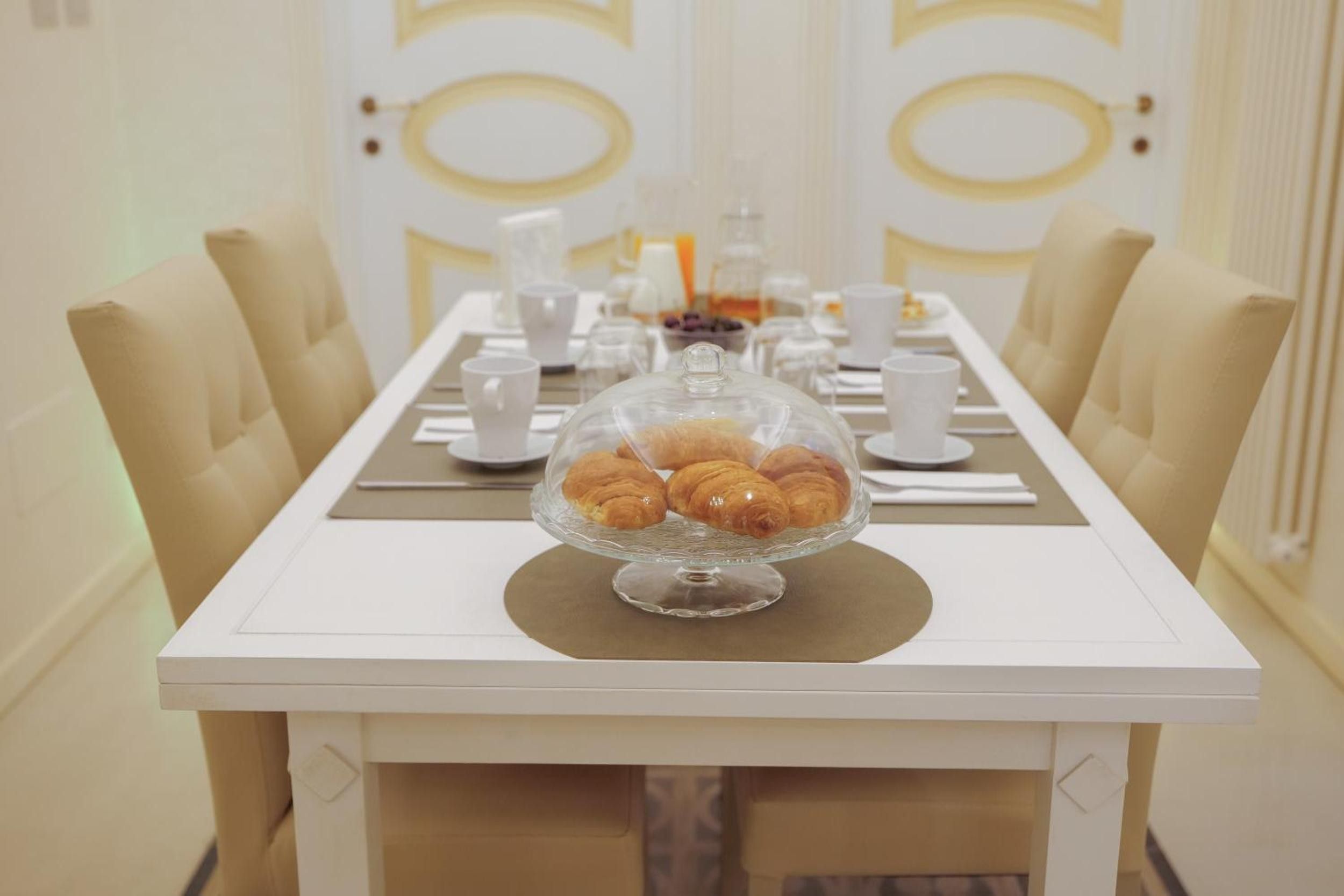 Une table à manger surmontée d’une cloche de verre contenant des pâtisseries, entourée de chaises blanches et recouverte d’une nappe blanche.