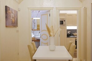 Un coin repas avec une table et des chaises blanches, un vase contenant des plantes séchées et une vue sur la cuisine.