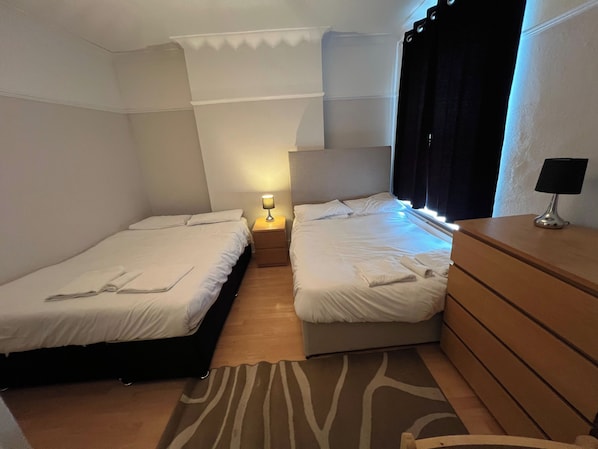 A hotel room with two single beds, a wooden dresser, a nightstand with a lamp, and a window with curtains.