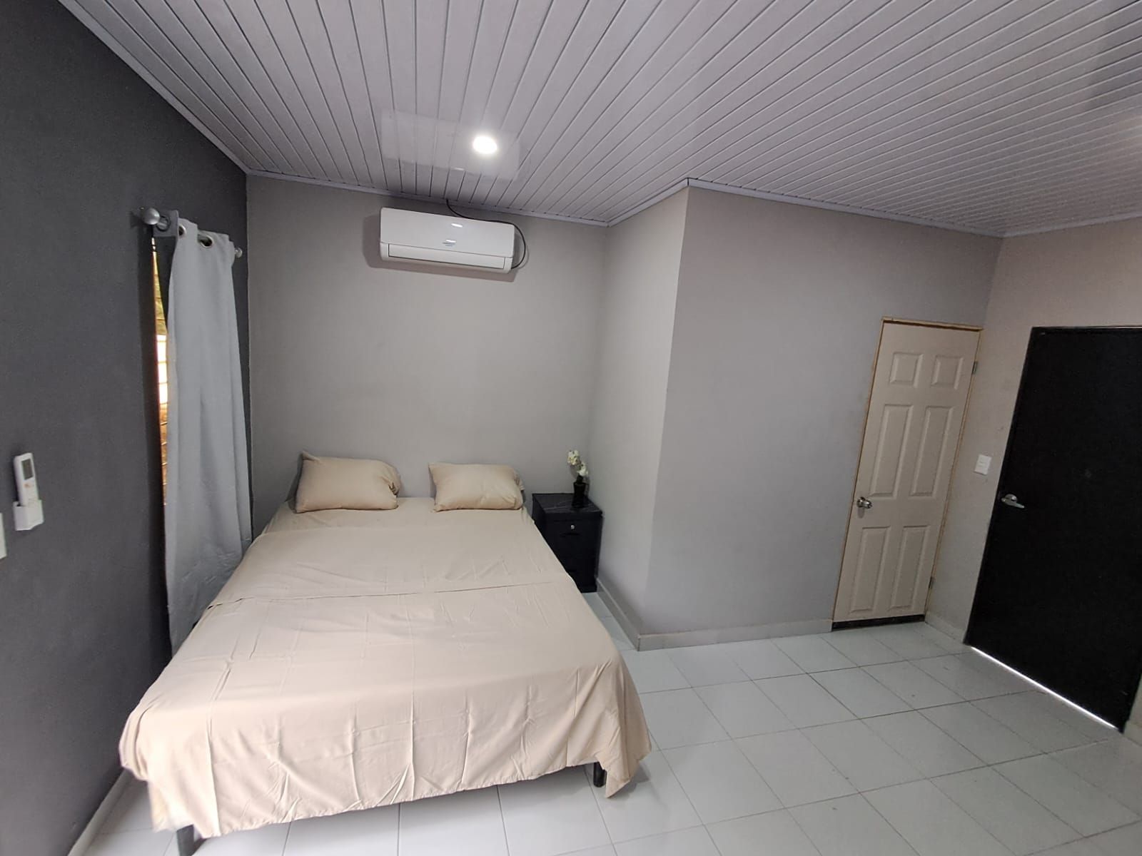Ein Schlafzimmer mit einem Bett, einem Nachttisch, einer Klimaanlage, einer Tür und einem gefliesten Boden.