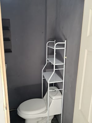 Un baño pequeño con un inodoro blanco, una estantería metálica blanca y una pared gris.