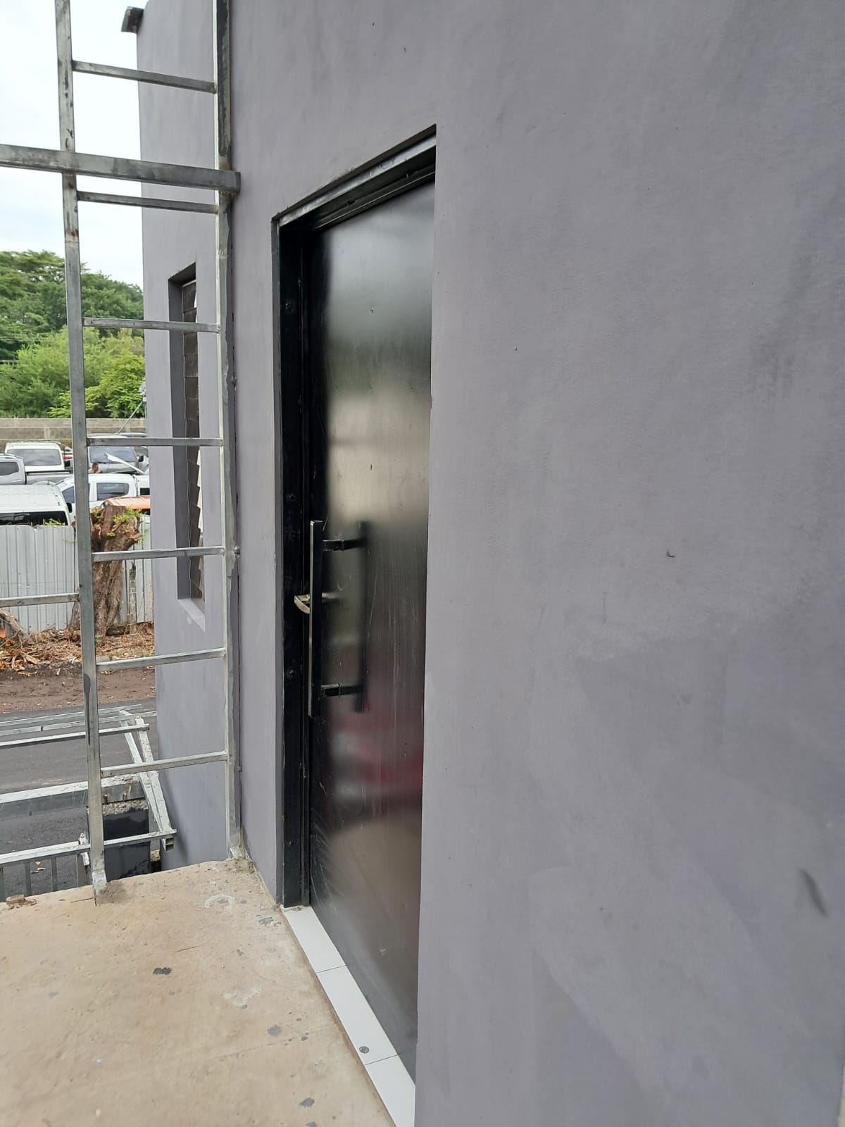 Fachada de un edificio con una escalera metálica, una puerta negra y un estacionamiento al fondo.