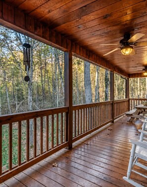 Eine überdachte Holzterrasse mit Deckenventilator, einer Hängeleuchte und Blick auf einen Wald.