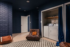 A laundry room with a washing machine, dryer, and a brown leather chair with a cushion. - Ruckle Retreat | Hot Tub + Sports Den + Putt Putt (Indianapolis)
