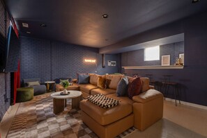 A modern living room with a sectional sofa, a coffee table, and a checkered throw. - Ruckle Retreat | Hot Tub + Sports Den + Putt Putt (Indianapolis)