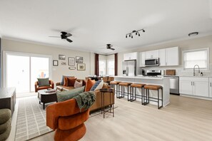 A modern living room with a sofa, coffee table, and a dining area with chairs. There is a kitchen with white cabinets, a microwave, and a refrigerator. - Ruckle Retreat | Hot Tub + Sports Den + Putt Putt (Indianapolis)