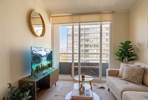 A living room with a flat-screen TV, a round mirror, a sofa, a coffee table, and a sliding glass door leading to a balcony. - Domingo Santa Maria by Domus Travels (Santiago)