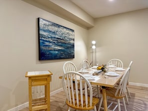 A dining area with a table set for four, white chairs, a wooden side table, and a large abstract painting on the wall. - Cedar Grove Residence (Surrey)
