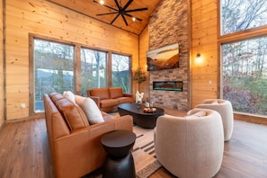 A cozy living room with a stone fireplace, leather sofas, and large windows offering a view of the outdoors. - HotTub/Theater/FirePit/GameRoom/5KingSuites! (Sevierville)