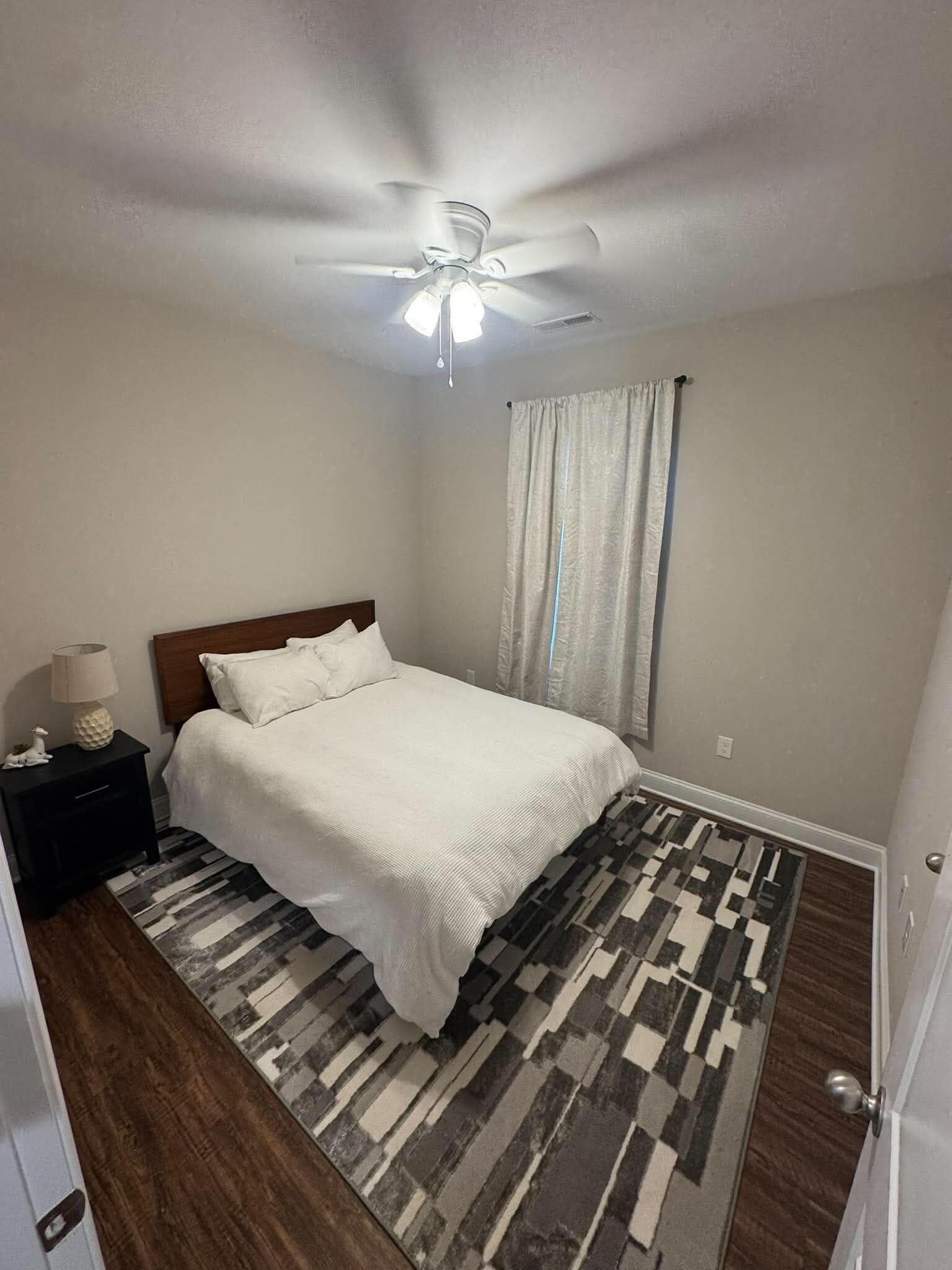 A bedroom with a bed, a nightstand, a ceiling fan, and a patterned rug.