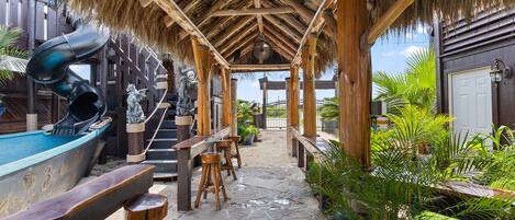 An outdoor bar area with a thatched roof, wooden stools, and a pool table.