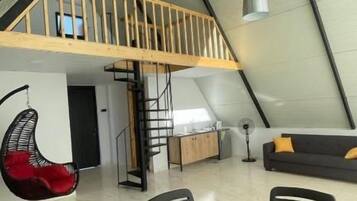 A spacious loft with a high ceiling, a dining area with a table and chairs, a red hanging chair, and a staircase leading to a lofted sleeping area.