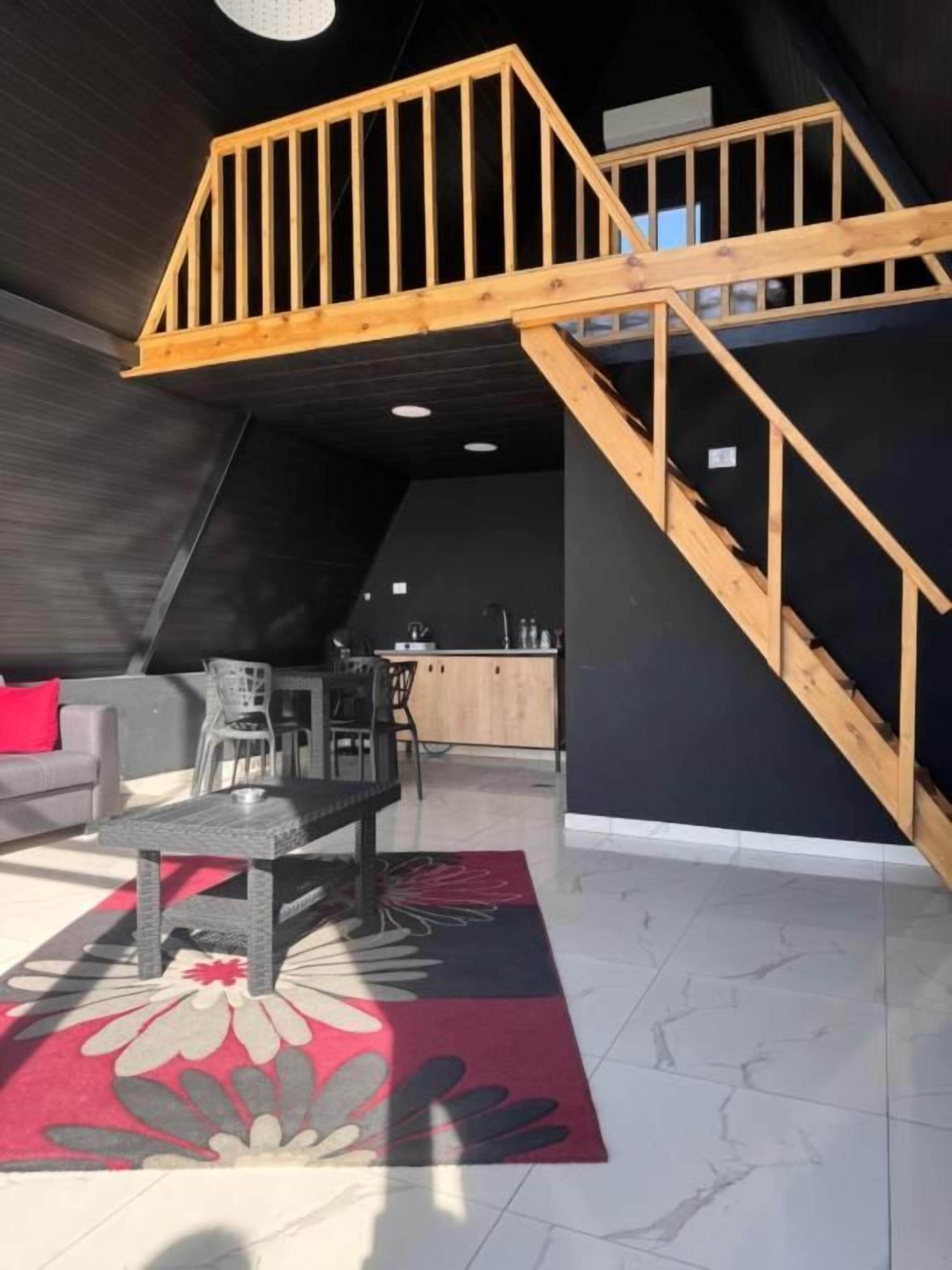 An interior with a wooden staircase, a dining area with chairs, a sofa, and a kitchen in the background.