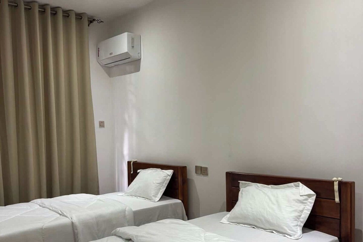 Habitació d'hotel amb dos llits individuals, un capçal de llit de fusta, una cortina beix i un aire condicionat fixat a la paret.