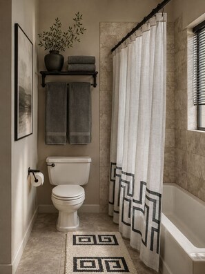 Een badkamer met een toilet, een handdoekrek met twee handdoeken, een plant in pot, een douchegordijn met een geometrisch patroon en een lijst met een foto aan de muur.