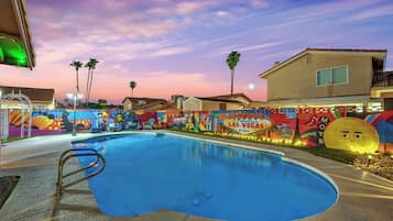 A swimming pool with a colorful mural in the background, a clear sky, and a building with a balcony.