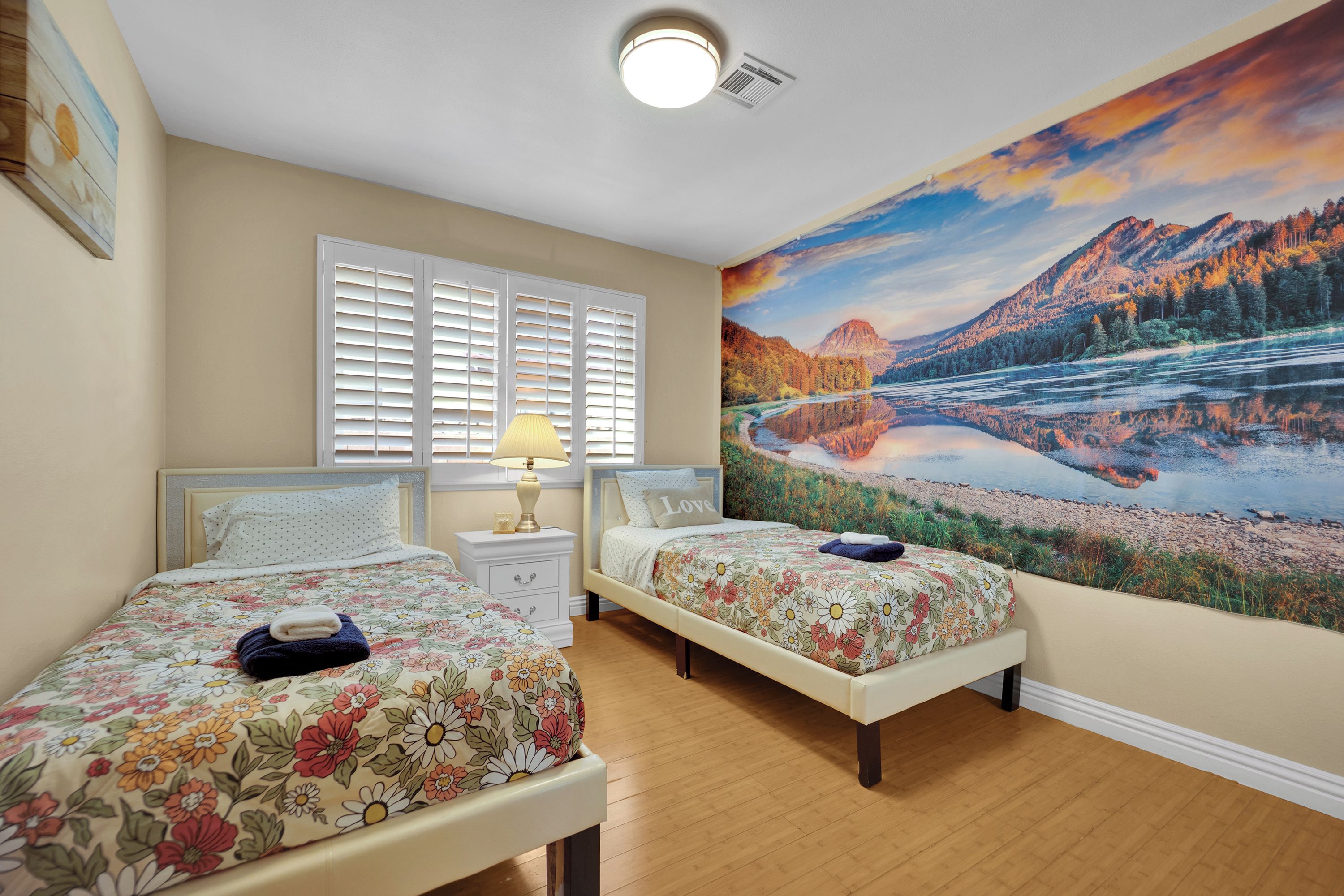 A bedroom with two beds, a nightstand, a lamp, and a large scenic wall mural.