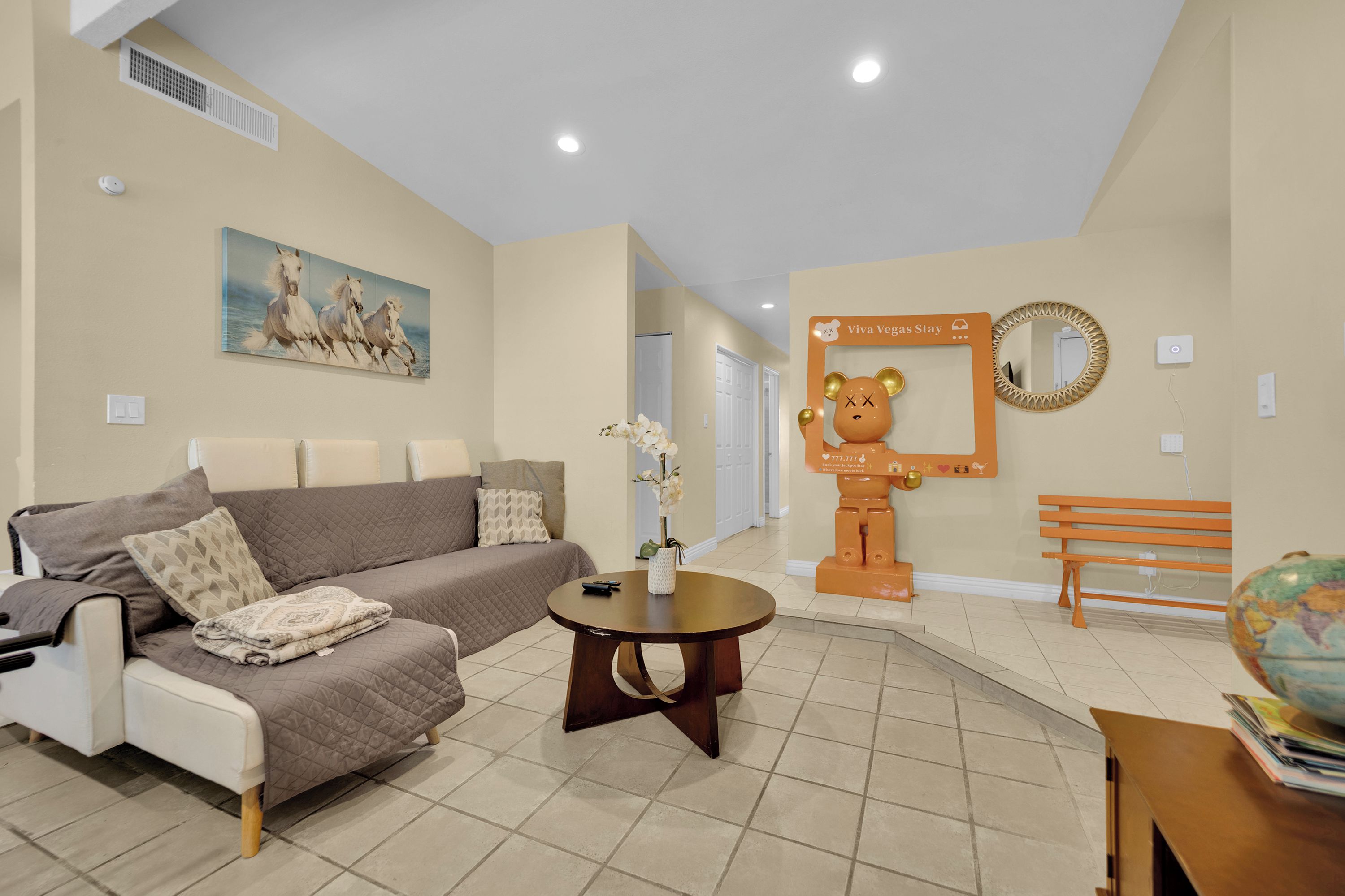 A living room with a sofa, a coffee table, a painting of horses, a bear-shaped sign, and a bench.
