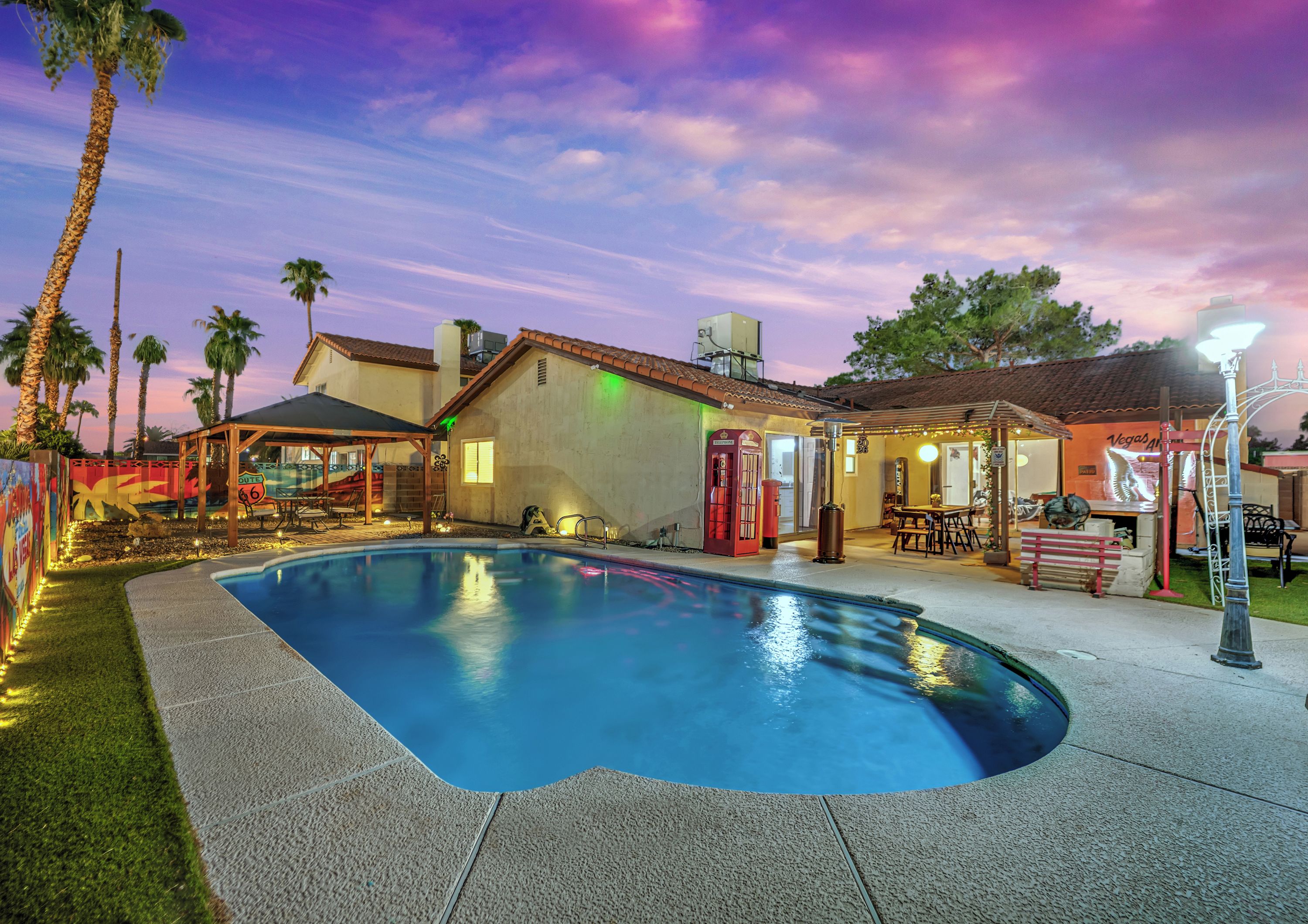 A swimming pool with a curved edge, surrounded by a patio area with outdoor seating and a small building with a red door.