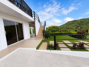 Una casa moderna con puerta de cristal, una escalera que lleva a un balcón, y un jardín cuidado con vistas a una colina verde.